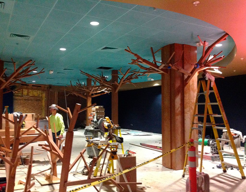 An in-progress photo of an indoor theme construction project, where COST engineers install artificial tree pillars with branches. The ceiling of the room is painted with theme elements, made to look like a sky.