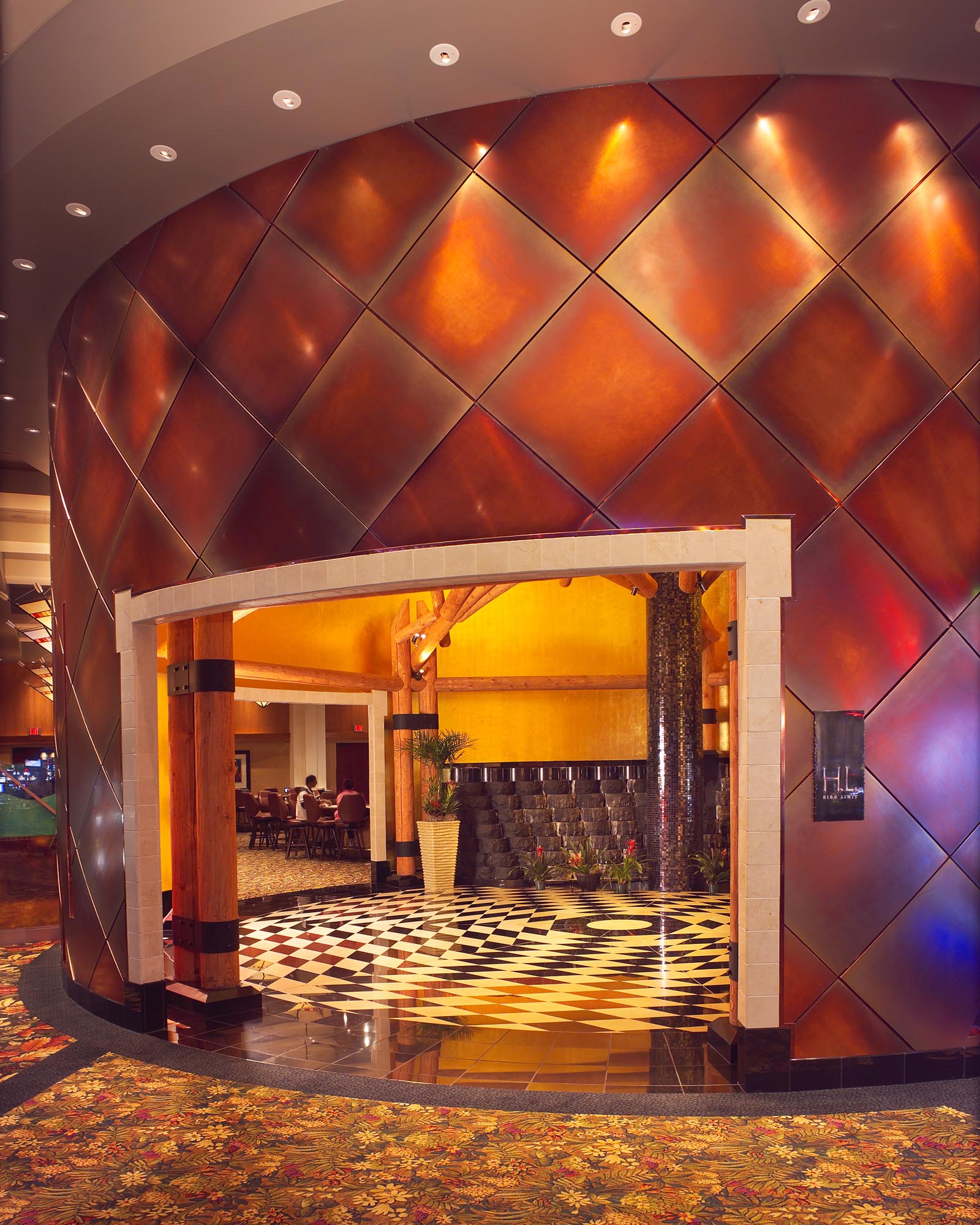 Four winds casino The entrance to this casino floor is separated by a large curved wall. The wall rises up from a patterned carpet and marble floor, and is covered in brass tiles that reflect the warm lighting.