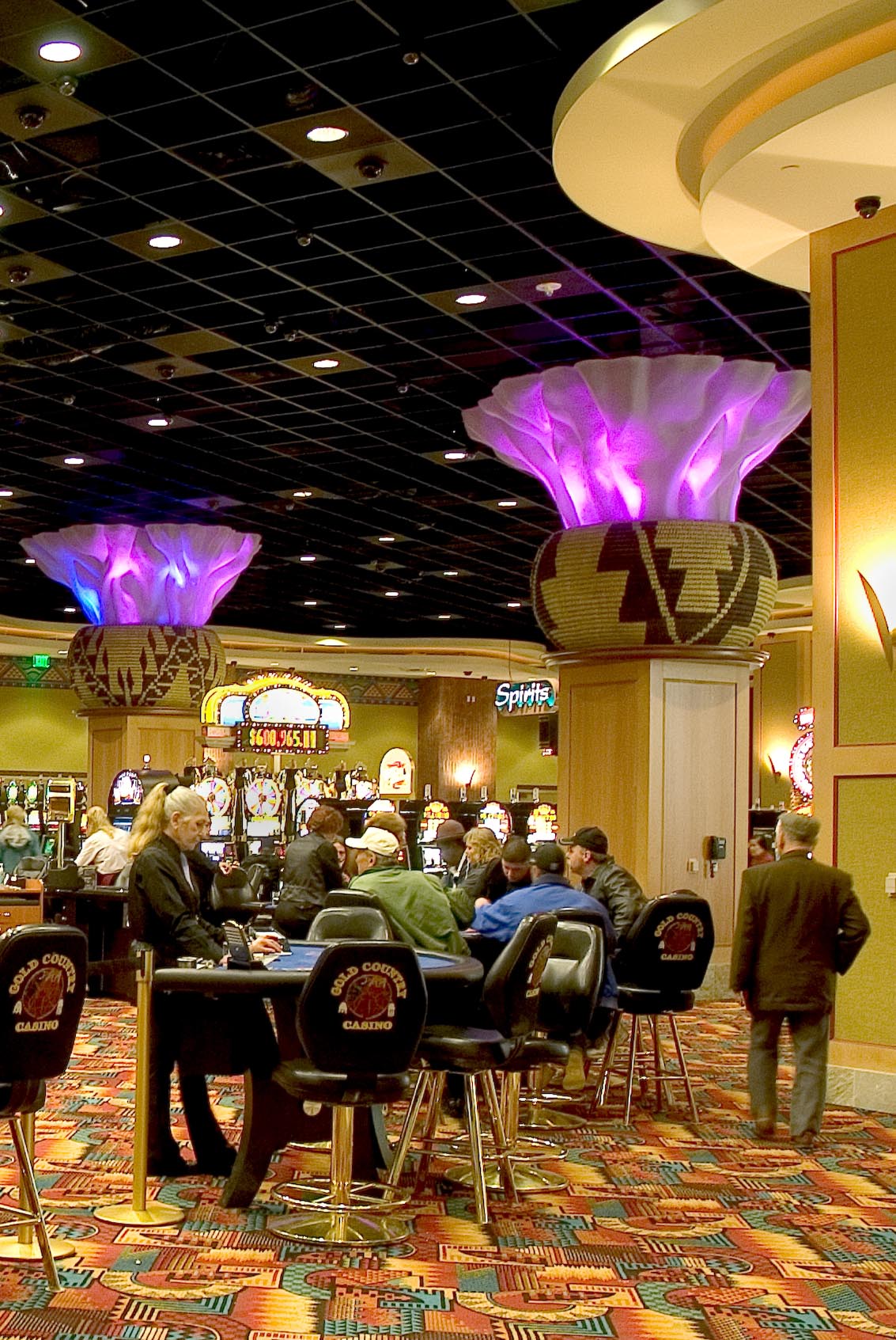 Goldcountry2 The casino floor features giant square pillars topped with Aztec patterened tiles and neon purple lights. Casino patrons visit blackjack tables and slot machines.