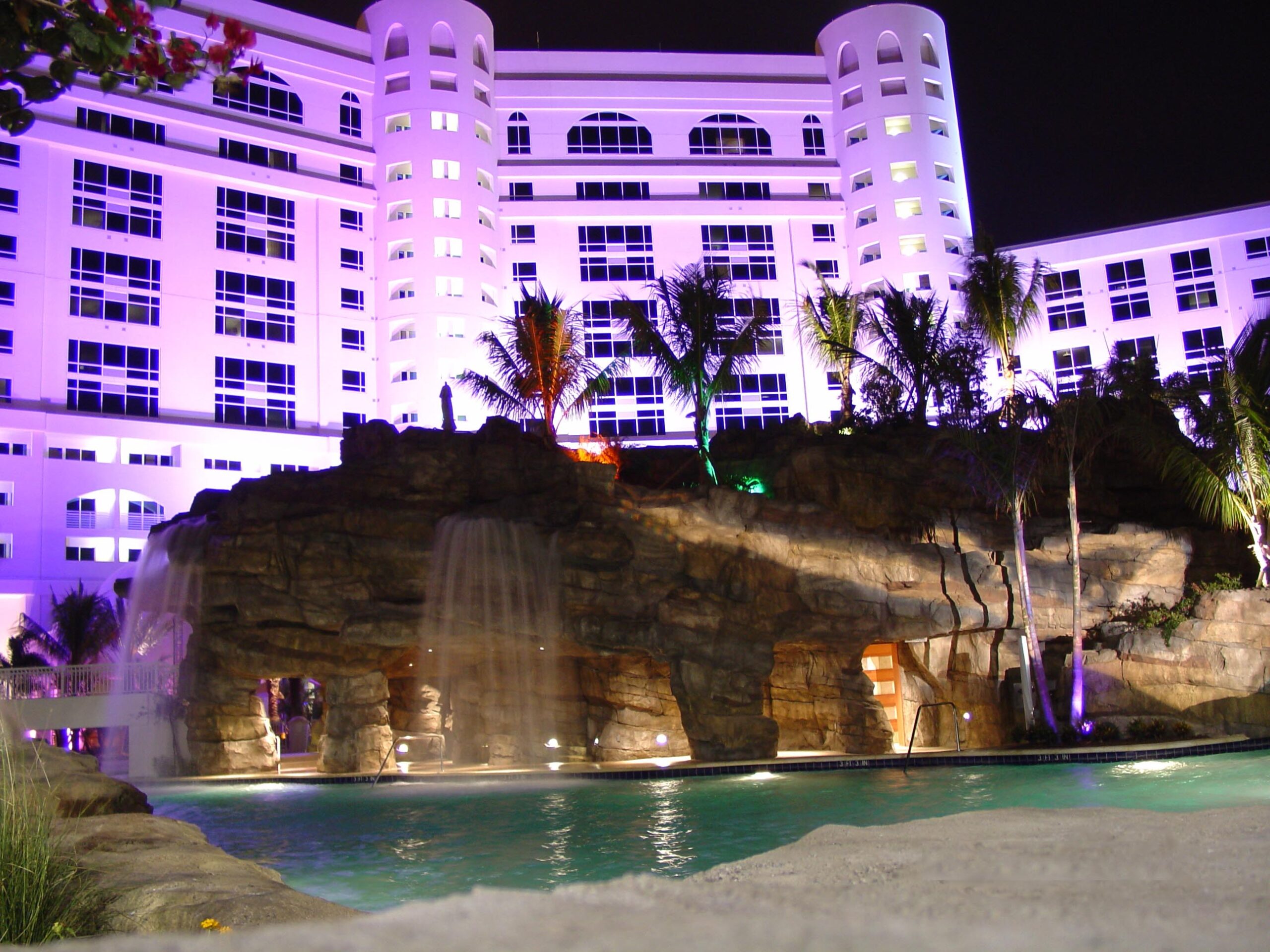 Hard rock night1 The exterior façade of the Hard Rock Hotel at night. The water feature in front of the hotel features waterfalls and a pool and mountain-like rockwork with trees and artificial vegetation. The hotel is lit with soft purple lighting.