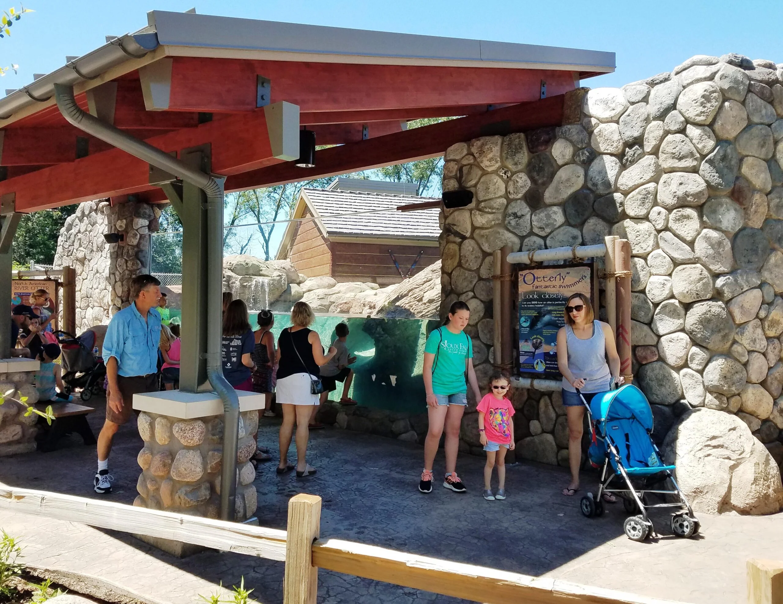 Mcz otters2 Zoo patrons gather around the exhibit window of the Minnesota Zoo otters exhibit. From this view, a large glass window is sheltered by a wood awning and artificial rock wall where patrons can look into the habitat.