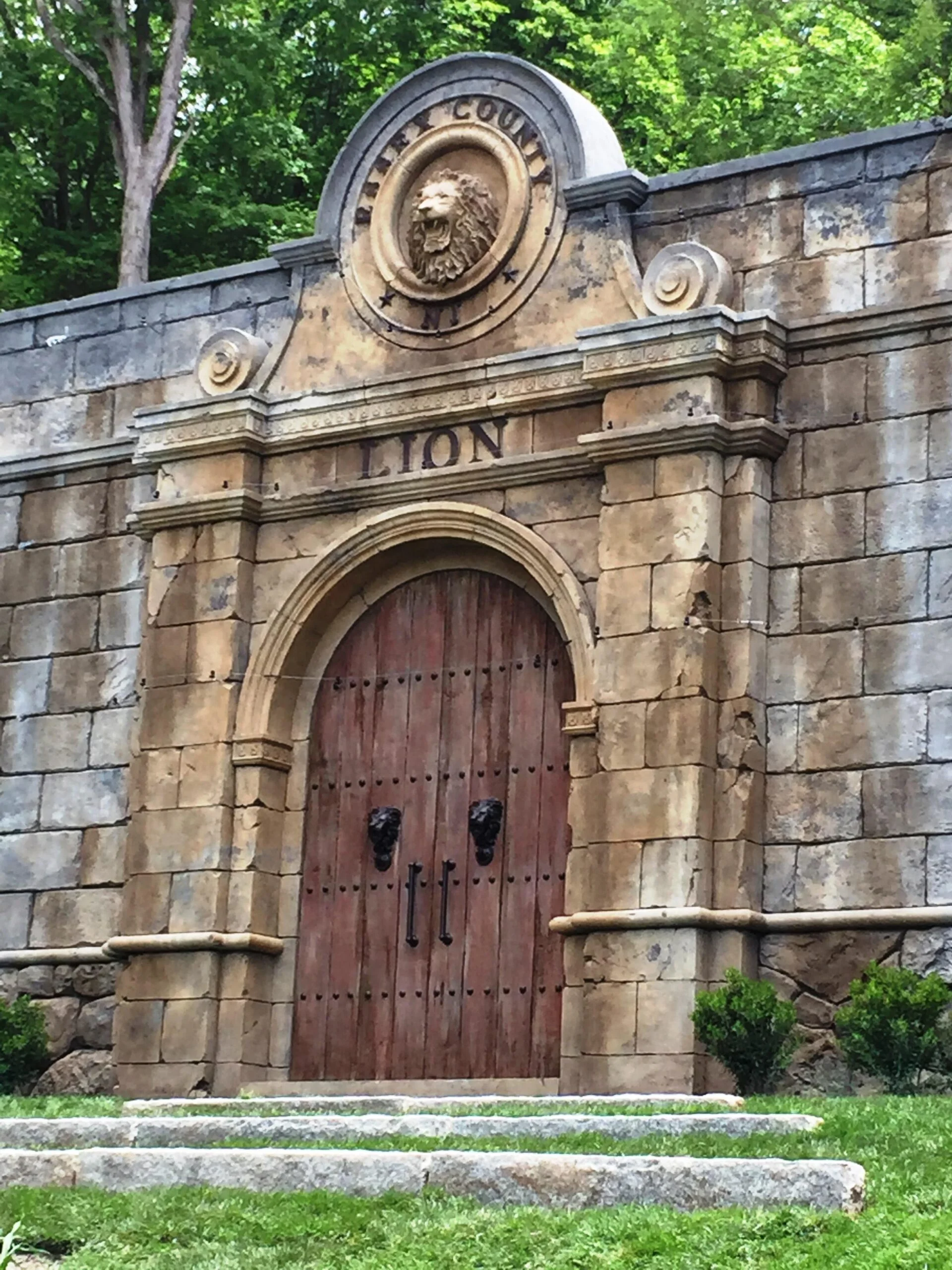 Turtle back zoo1 A grand stonework arch entrance to the Turtle Back Zoo lion habitat features a weathered stone wall and two grand wood doors. Above the doors, the word "LION" is carved into the artificial rockwork, and is topped with a bust statue of a lion.